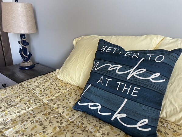 Una cama con ropa de cama floral, un almohadón con el texto "Better to wake at the lake" y una lámpara en una mesita de noche.