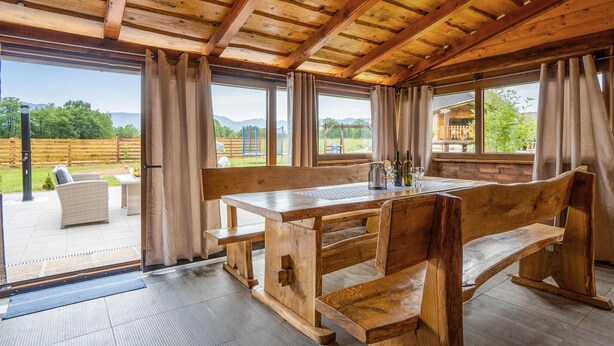 Un comedor de madera con banco y mesa, ventanas amplias y vista a un espacio exterior cercado.