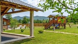 Una cabaña de madera con un porche cubierto, una estructura de juegos y un patio césped con montañas al fondo.