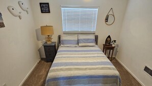 A bedroom with a bed, a ceiling fan, a window with blinds, a nightstand, a lamp, and wall decorations.