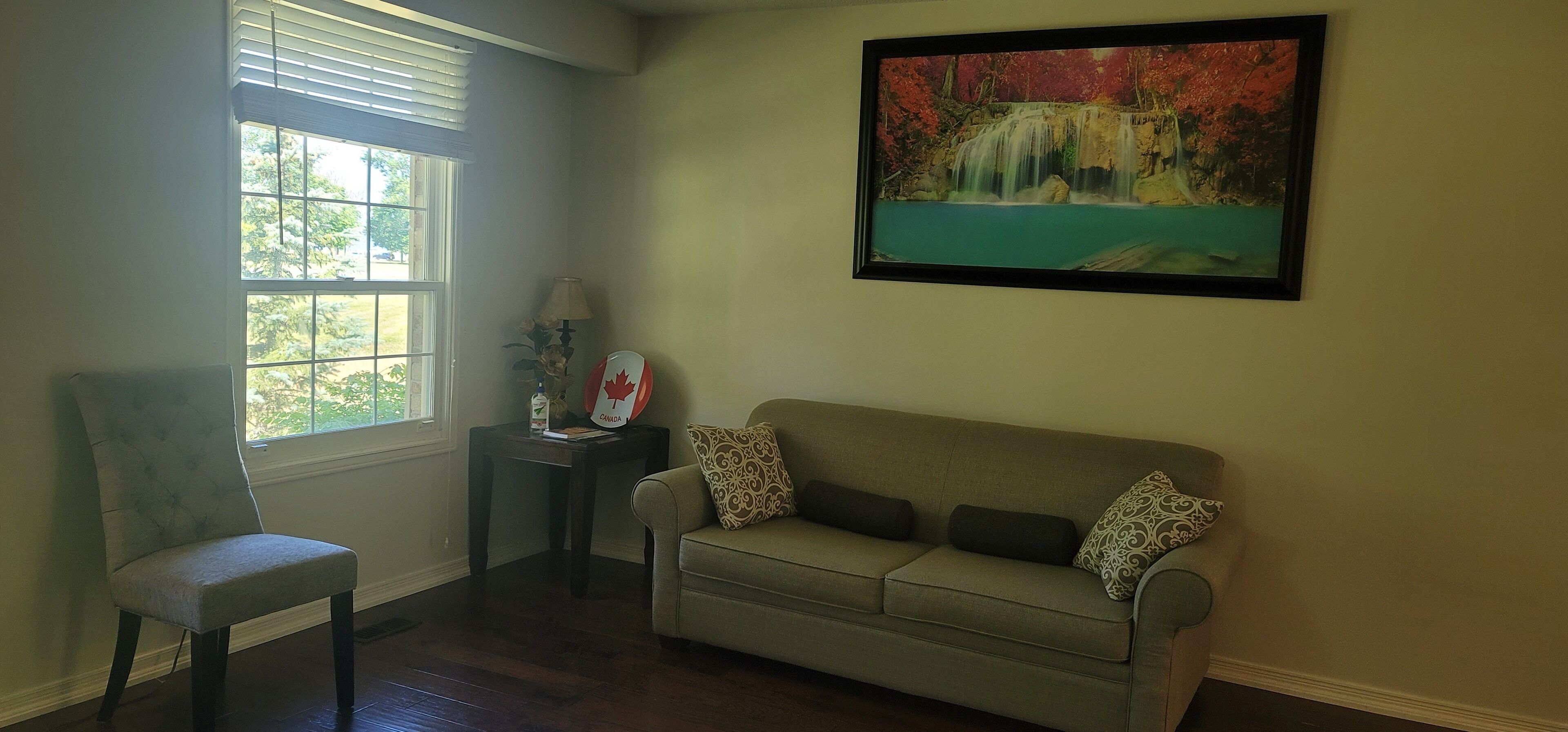 Un salón con un sofá color beige, una silla, una mesita con lámpara y un cuadro con una cascada.