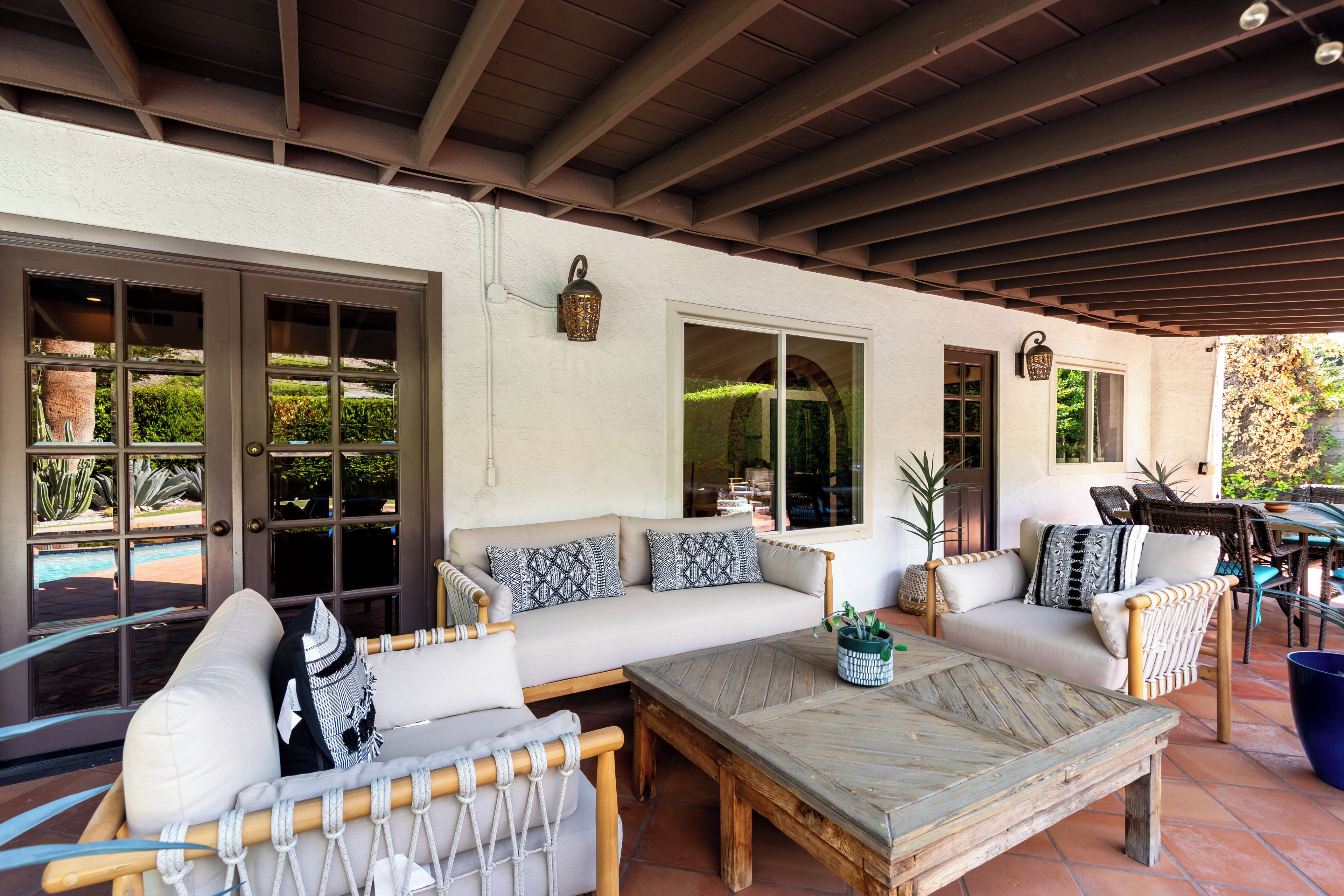 A covered patio area with a sofa set, a wooden table, and a view of the outdoors through large windows.