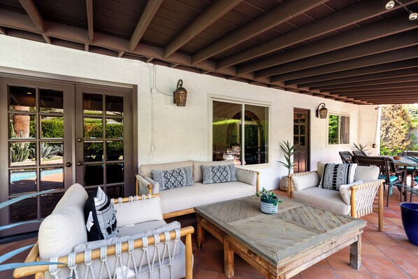 A covered patio area with a sofa set, a wooden table, and a view of the outdoors through large windows.