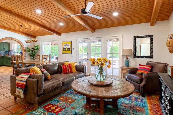 A spacious living room with a brown leather sofa, a round wooden coffee table, and a colorful rug. There is a ceiling fan, a mirror, and a door leading outside. - Casa Fierro: 3-bed Superior Spanish retreat in south Palm Springs (Palm Springs)