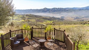 A wooden deck with wicker furniture and potted plants, overlooking a valley with mountains and a river. - Beautiful home in Periana (Periana)