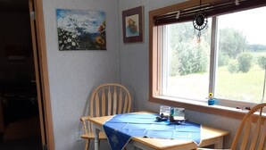 Una habitación con una mesa y sillas de madera, una ventana que da a zonas verdes y una puerta que conduce a otra habitación.