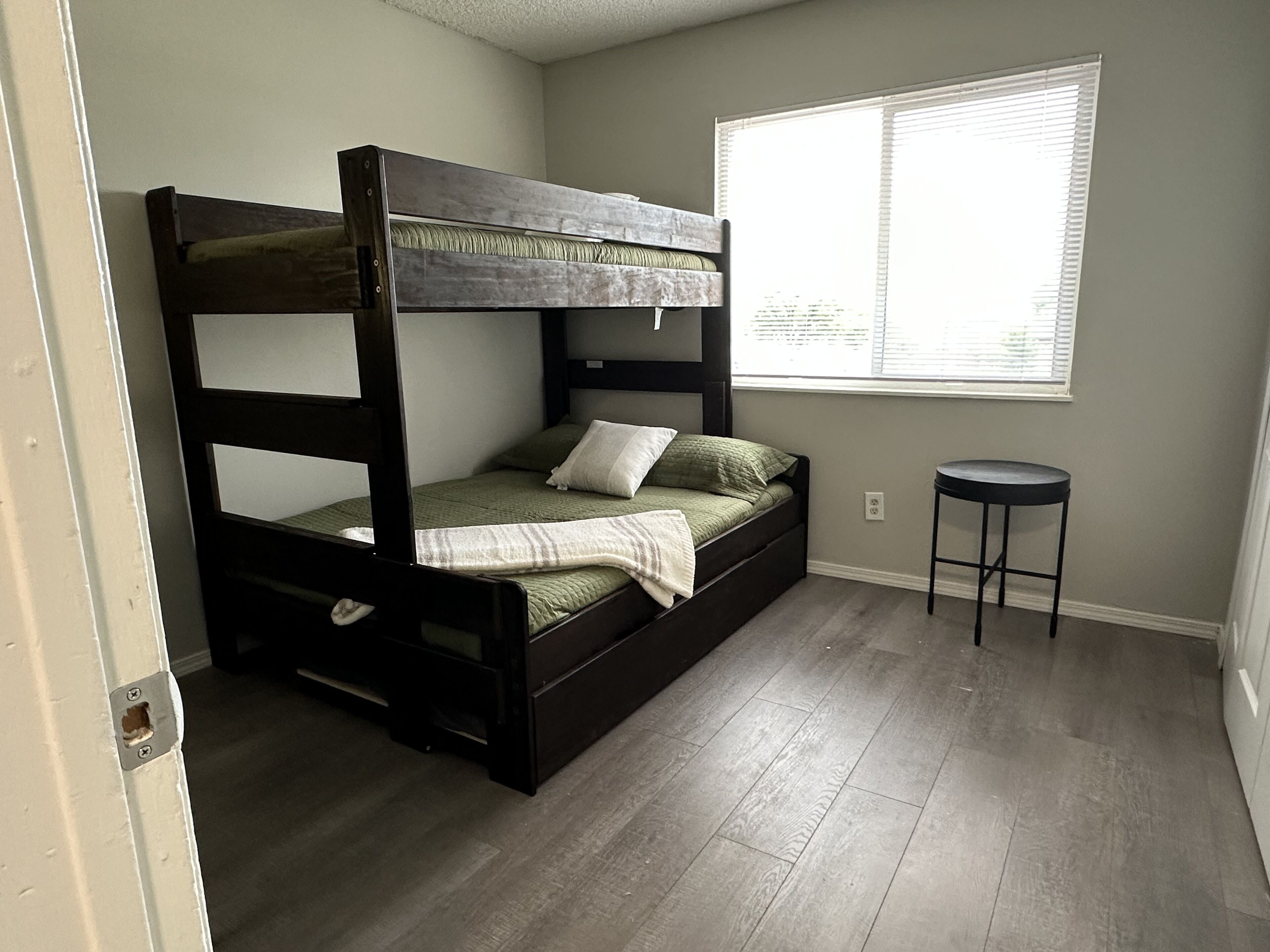 A bunk bed with a wooden frame, a small stool, and a window with blinds.