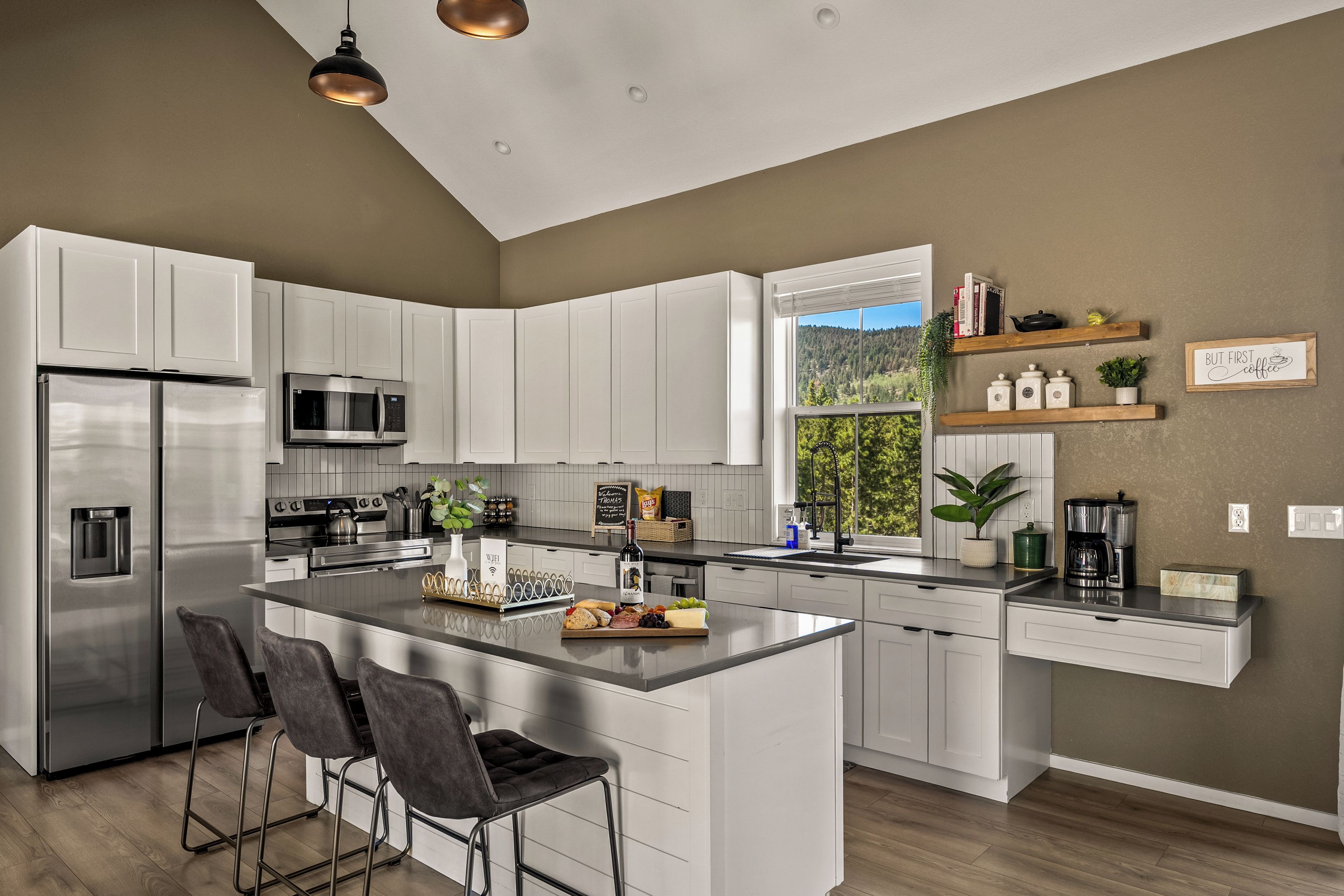 Una cocina moderna con armarios blancos, electrodomésticos de acero inoxidable, una isla central con taburetes y vistas a un bosque por una ventana.