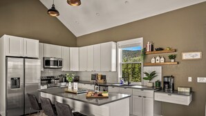 A modern kitchen with white cabinetry, stainless steel appliances, a central island with bar stools, and a view of a forest through a window.