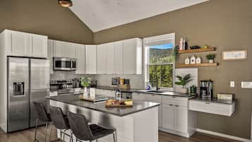 Una cocina moderna con armarios blancos, electrodomésticos de acero inoxidable, una isla central con taburetes y vistas a un bosque por una ventana.