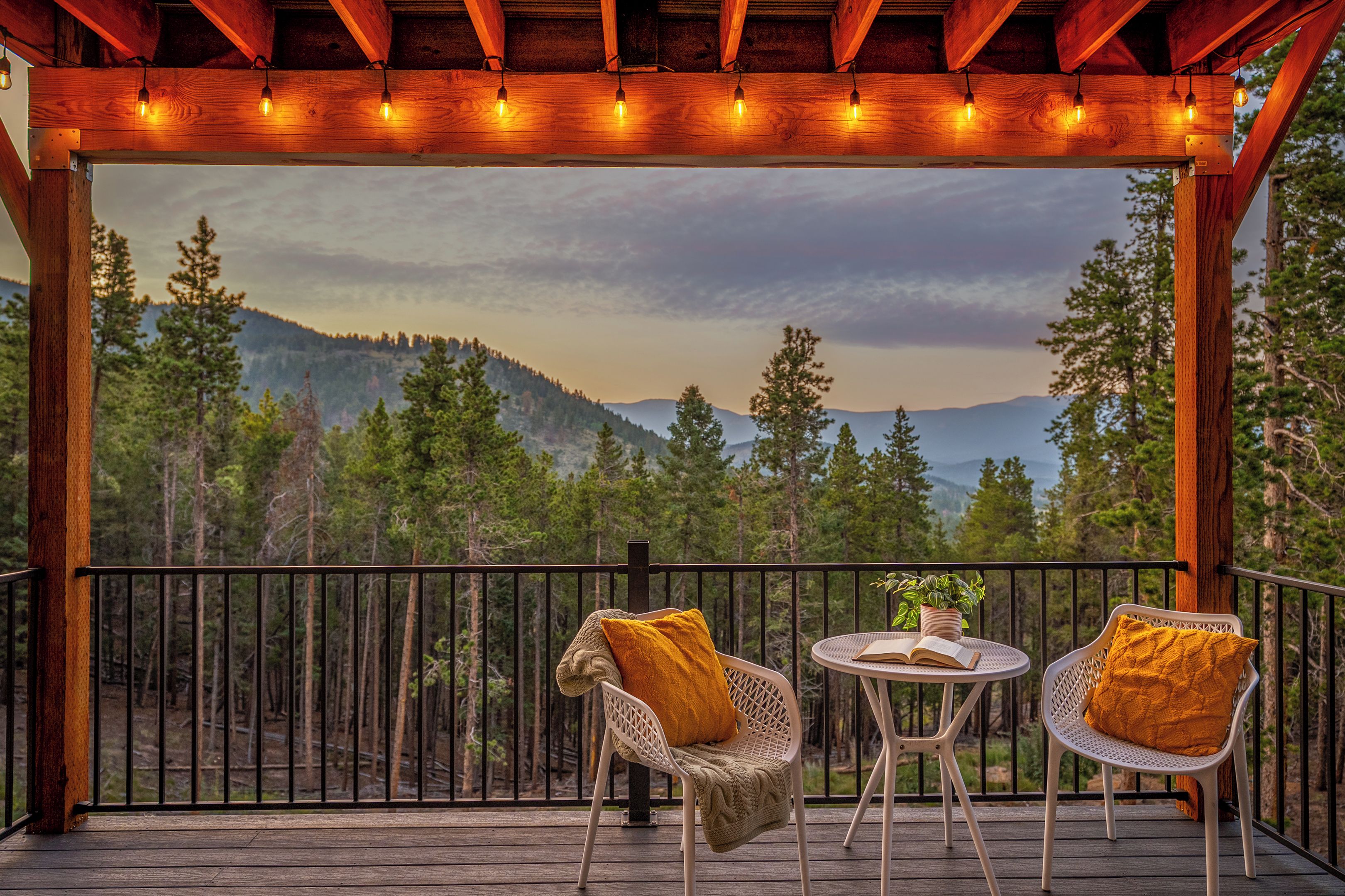 Eine Holzterrasse mit zwei Korbstühlen und einem kleinen Tisch, mit Blick auf einen bewaldeten Gebirgszug.