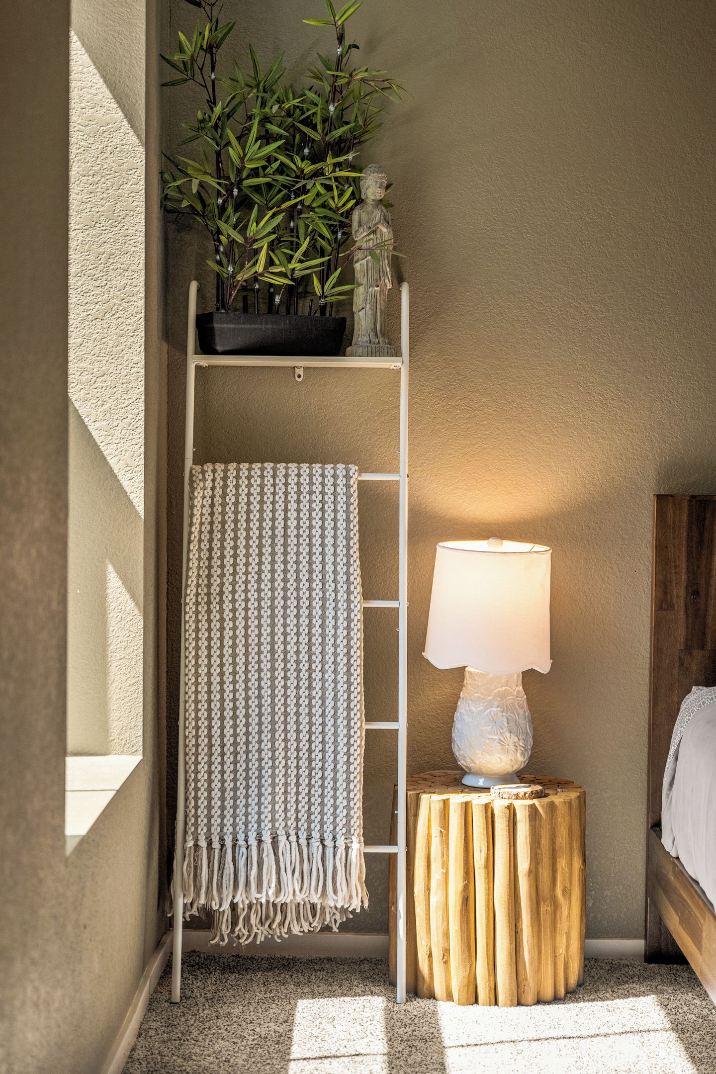A corner room with a wooden ladder shelf holding a plant, a hanging towel, a lamp on a wooden stool, and a bed.