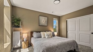A bedroom with a bed, a nightstand, a wall-mounted lamp, a decorative wall piece, a window, and a door.