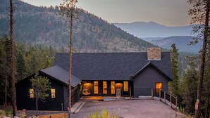 A modern building with a dark exterior, a paved driveway leading to the entrance, and a view of a forested mountain range in the background.