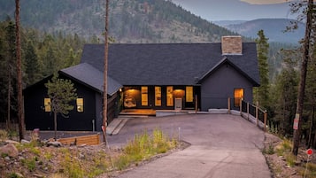 A modern building with a dark exterior, a paved driveway leading to the entrance, and a view of a forested mountain range in the background.
