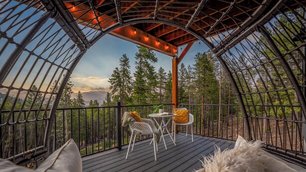 A cozy outdoor seating area with a table and chairs, surrounded by a wooden deck and metal railings, overlooking a forested landscape.