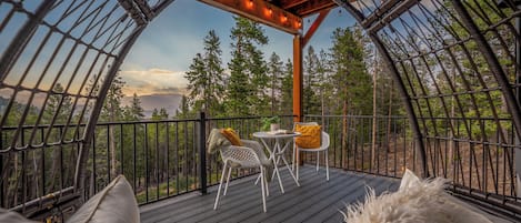 A cozy outdoor seating area with a table and chairs, surrounded by a wooden deck and metal railings, overlooking a forested landscape.