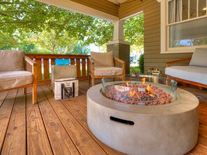 Una terraza de madera con mobiliario exterior, un brasero de vidrio y vistas a árboles.