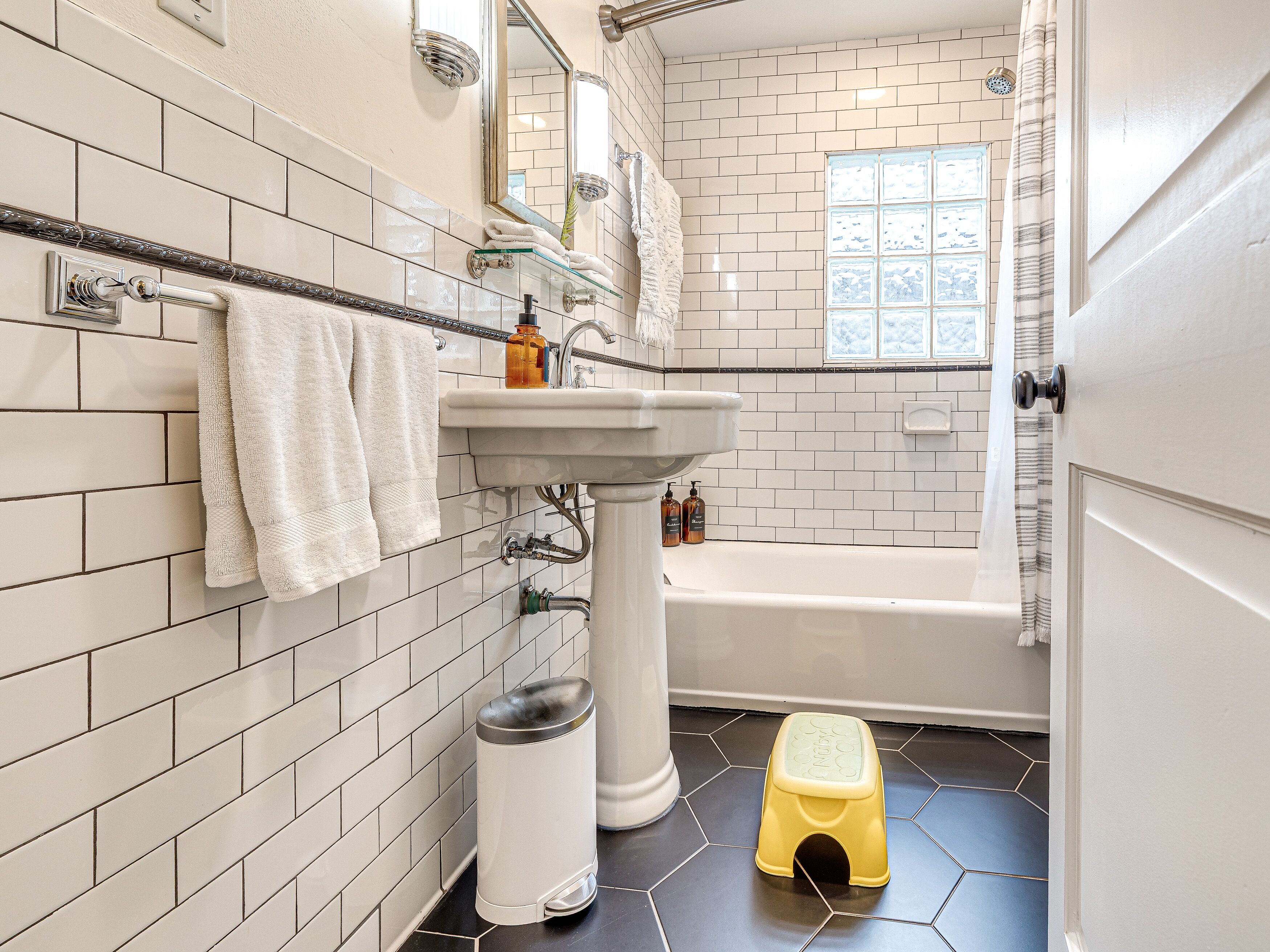 Une salle de bain avec des carreaux de céramique blancs rectangulaires, un lavabo blanc, une baignoire et une fenêtre donnant sur un beau paysage.