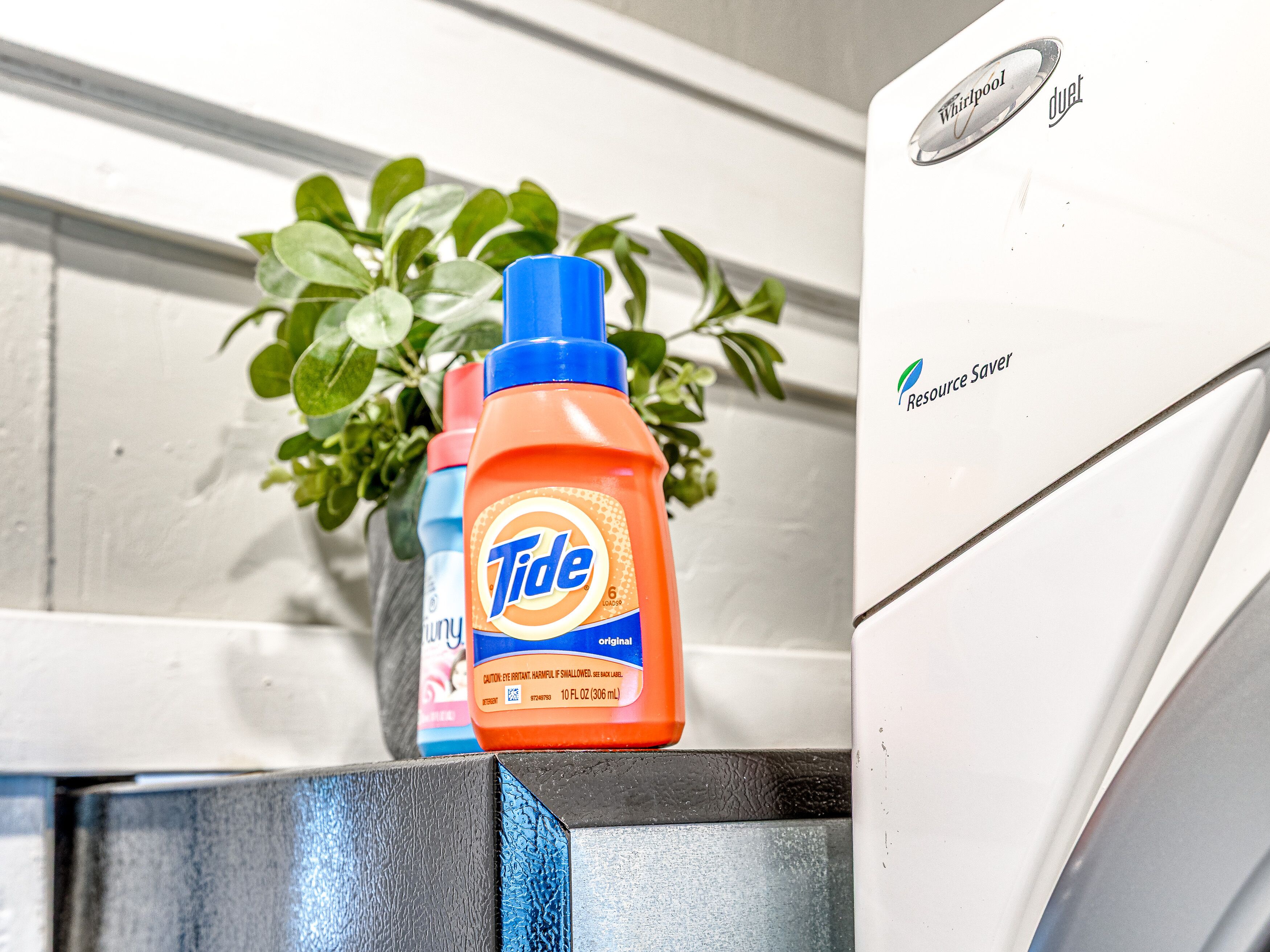 A Tide detergent bottle on a metal surface with a Whirlpool refrigerator in the background.
