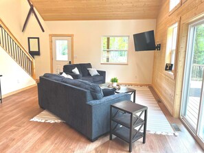 Sala de estar moderna com um sofá cinza escuro, uma mesinha de centro e uma TV fixada na parede. Janelas oferecem iluminação natural e vista para o exterior.