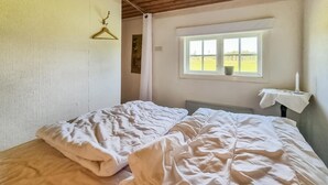 Een kamer met een bed, een raam met uitzicht op een groen veld, een hanglamp en een tafel met een kaars.