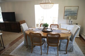 Uma área de refeições com uma mesa redonda, seis cadeiras de vime, um lustre e uma televisão ao fundo.