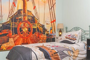 Un dormitorio con un tapiz grande con motivos marinos, una cama con cubrecama de temática náutica, cabecera metálica verde y una mesita de noche con lámpara.