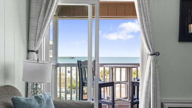 A room with a view of the ocean, a balcony with outdoor furniture, and a window with curtains.