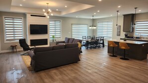 Ein modernes Wohnzimmer mit großzügigem Sofa, einem Essbereich mit Tisch und Stühlen, einer Küche mit Bar und einem an der Wand befestigten Fernseher.