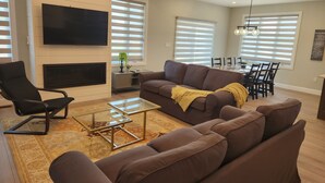 Ein modernes Wohnzimmer mit einem großen Flachbildfernseher, einem braunen Sofa, einem Glastisch und einem Essbereich mit Tisch und Stühlen.
