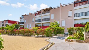 A row of multi-story residential buildings with balconies, a sandy courtyard, and various plants and shrubs.