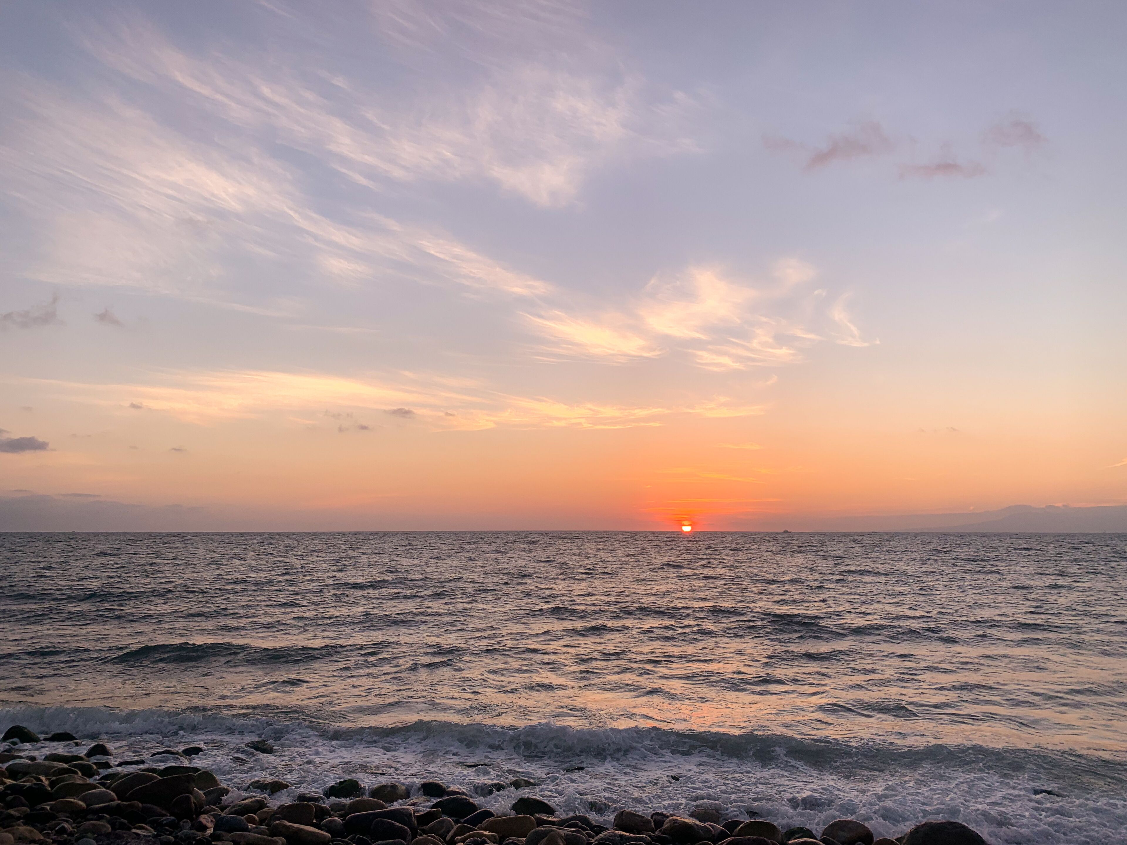 A sunset over the ocean with waves crashing onto a rocky shore.