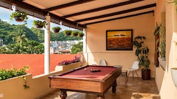 Una mesa de billar en una habitación con vistas a un paisaje urbano y zonas verdes, un cuadro en la pared y plantas en macetas.