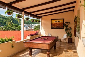 Una mesa de billar en una habitación con vistas a un paisaje urbano y zonas verdes, un cuadro en la pared y plantas en macetas.