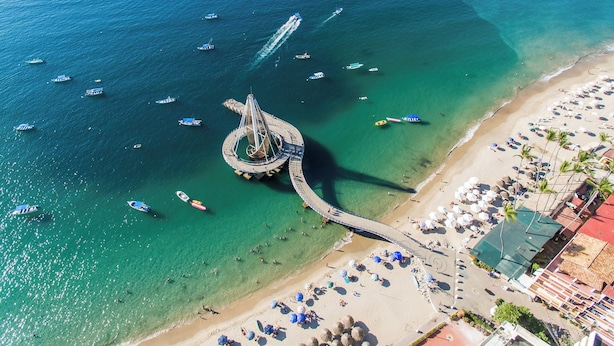 An aerial view of a coastal area with a central pier, boats, and a beach with umbrellas and people. - HOTEL PUEBLITO VALLARTA (Puerto Vallarta)