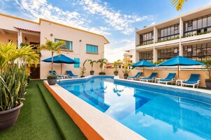 A swimming pool with lounge chairs and umbrellas, surrounded by buildings and potted plants.