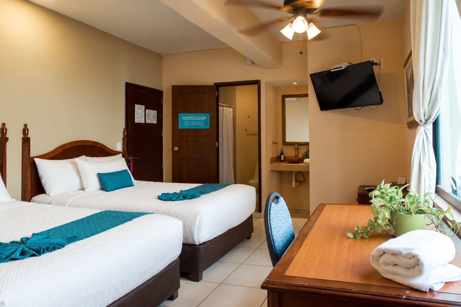 A hotel room with two beds, a wooden desk, a television, a bathroom, and a window with curtains.