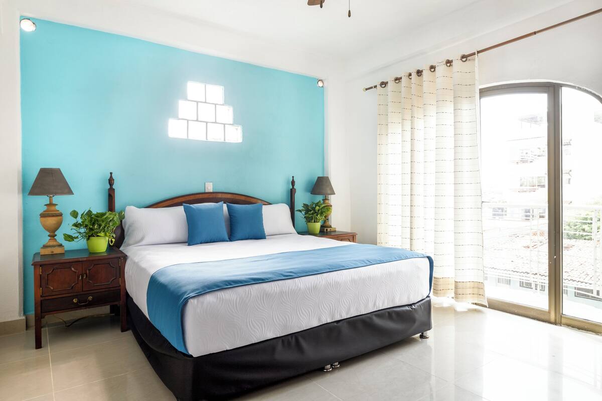 A bedroom with a bed, two nightstands, a window with curtains, and a view of buildings outside.