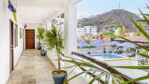 A balcony with a pool, potted plants, and a view of a hill with buildings. - HOTEL PUEBLITO VALLARTA (Puerto Vallarta)