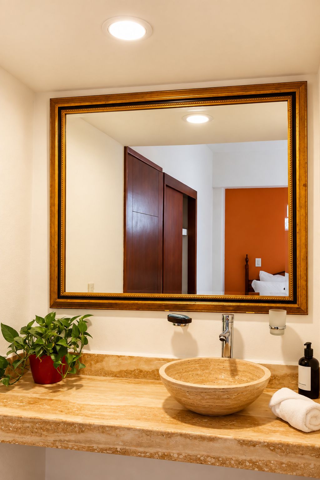 Un baño con tocador de madera, lavabo, espejo con marco dorado, planta en maceta y toalla.