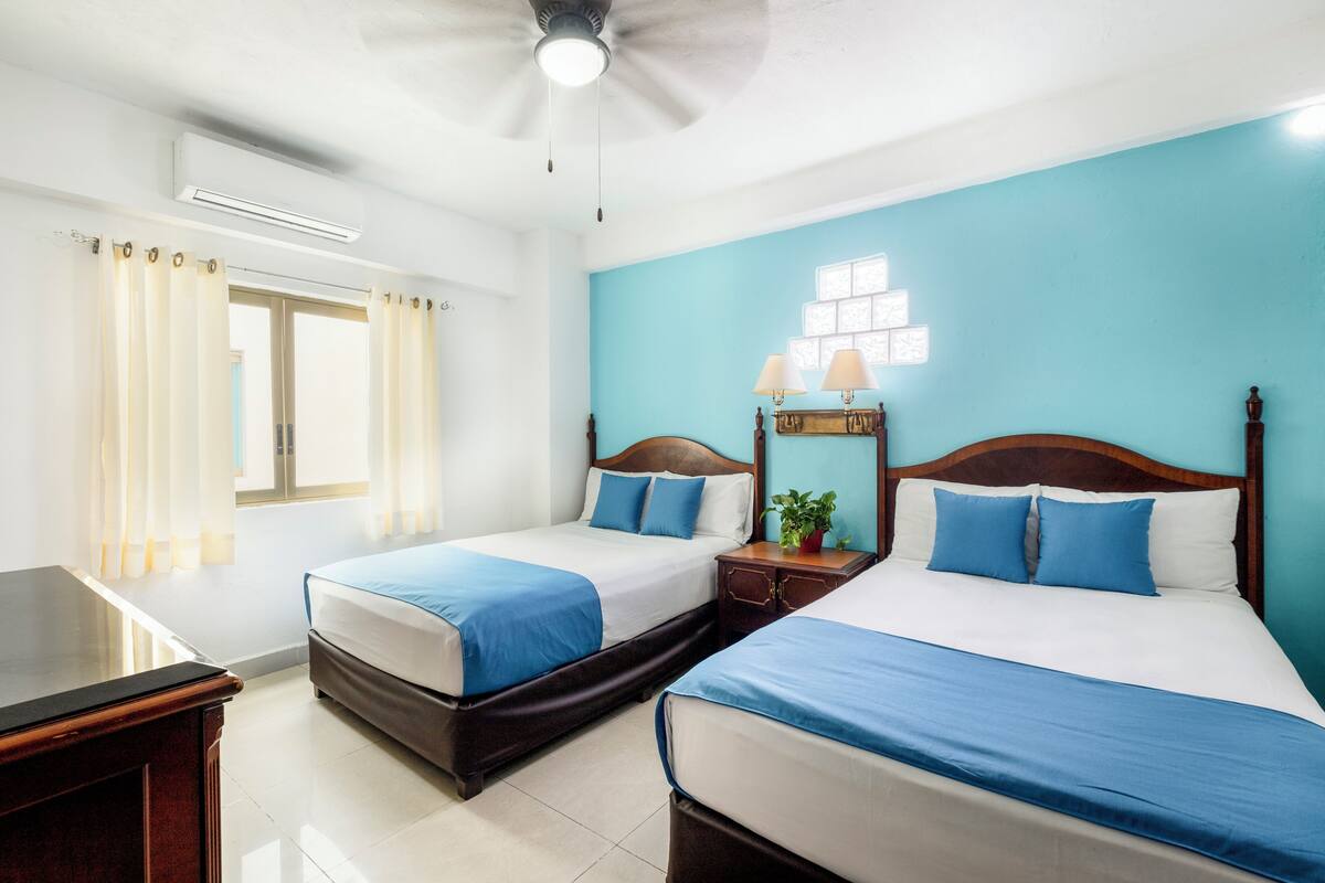 A hotel room with two beds, a desk, a ceiling fan, and a window with curtains.