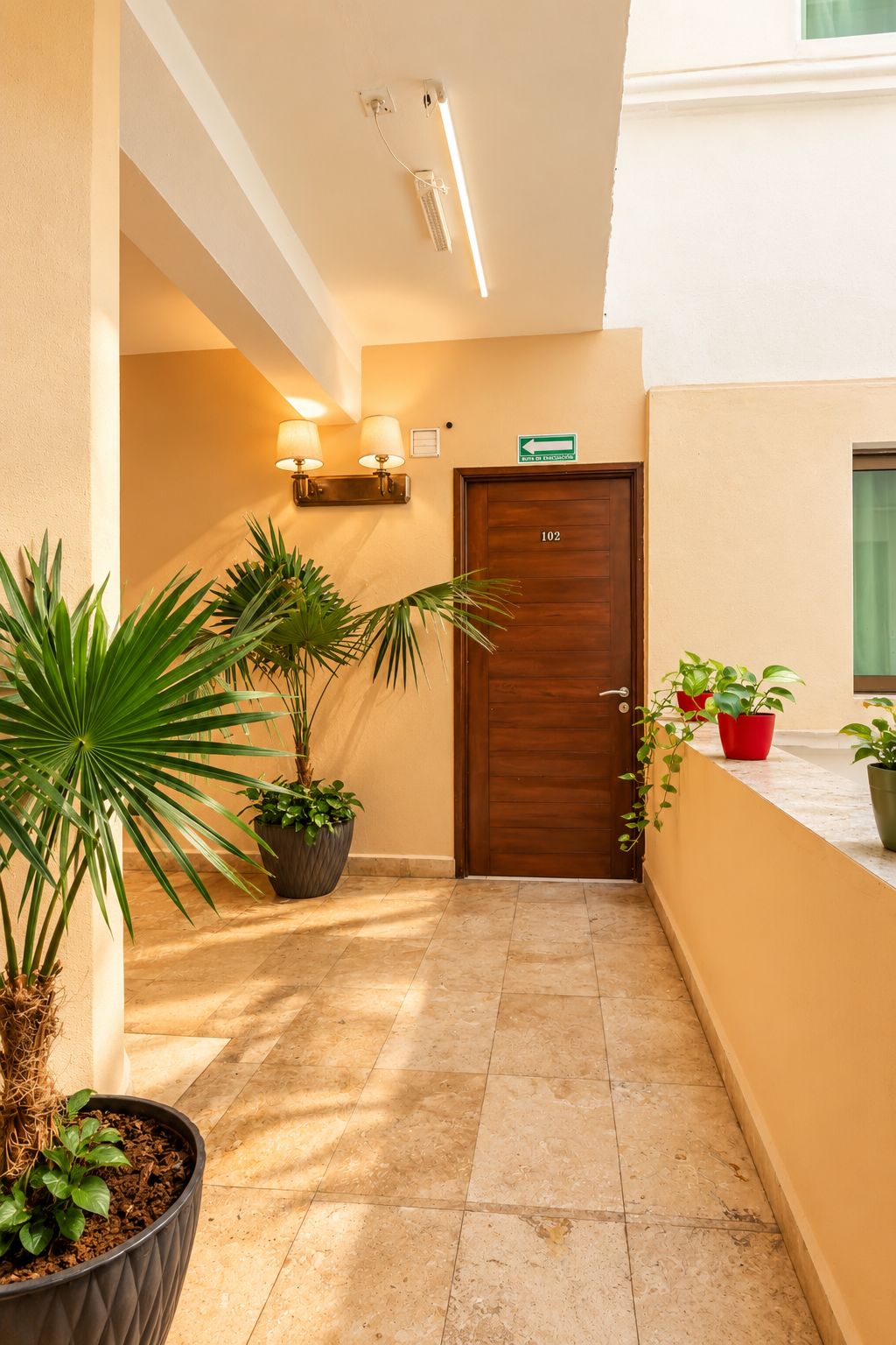Pasillo de hotel con puerta de madera con el número '102', plantas en macetas y lámparas fijadas a la pared.
