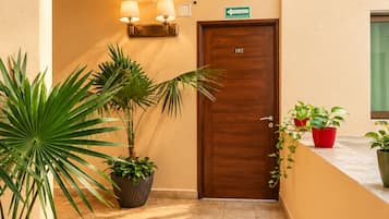 Pasillo de hotel con puerta de madera con el número '102', plantas en macetas y lámparas fijadas a la pared.