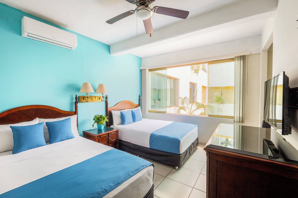 A hotel room with two beds, a ceiling fan, an air conditioning unit, a TV, and a large window with a view of the outdoors.