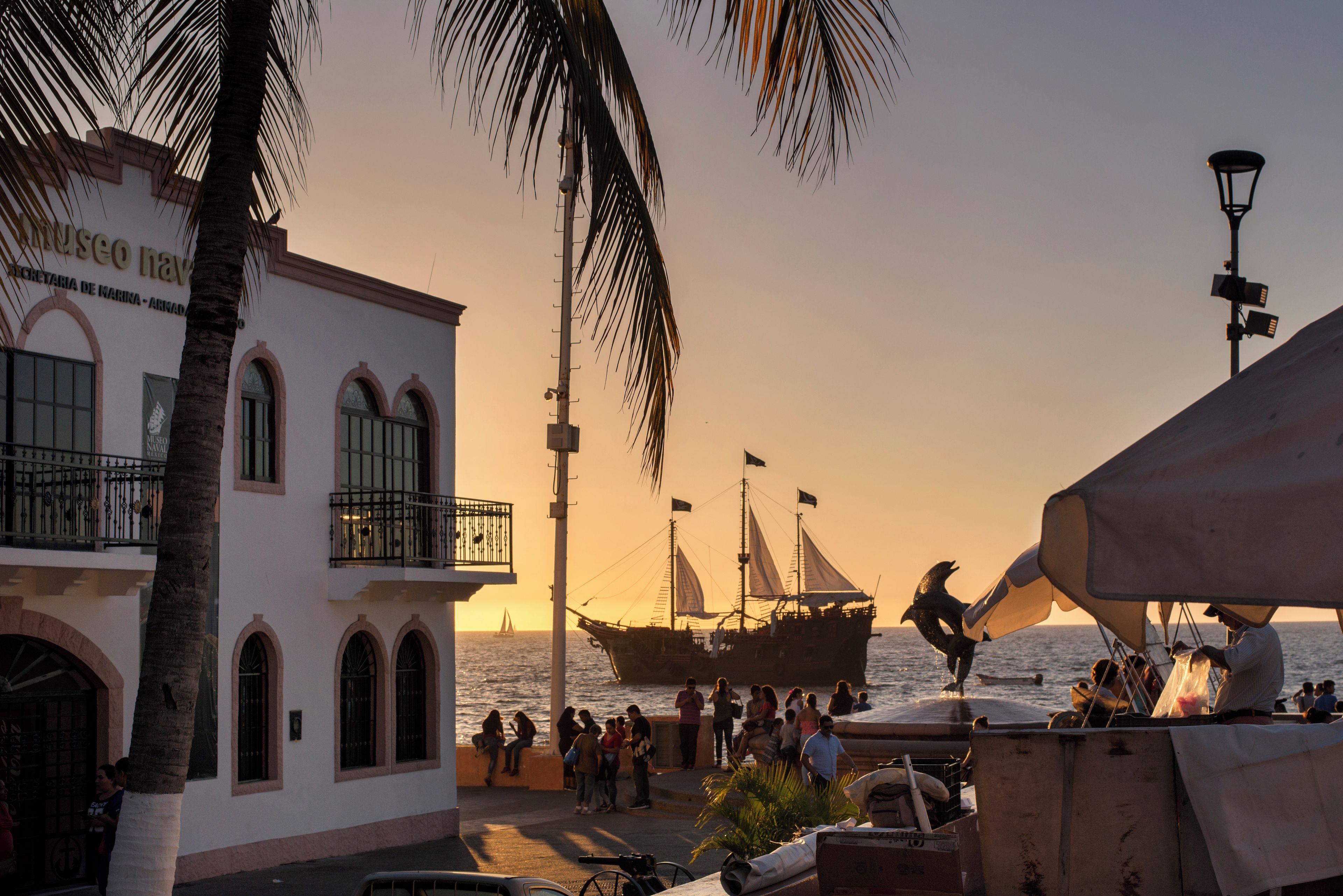 A coastal building with arched windows and a balcony, a sailboat in the background, and people gathered by the waterfront.