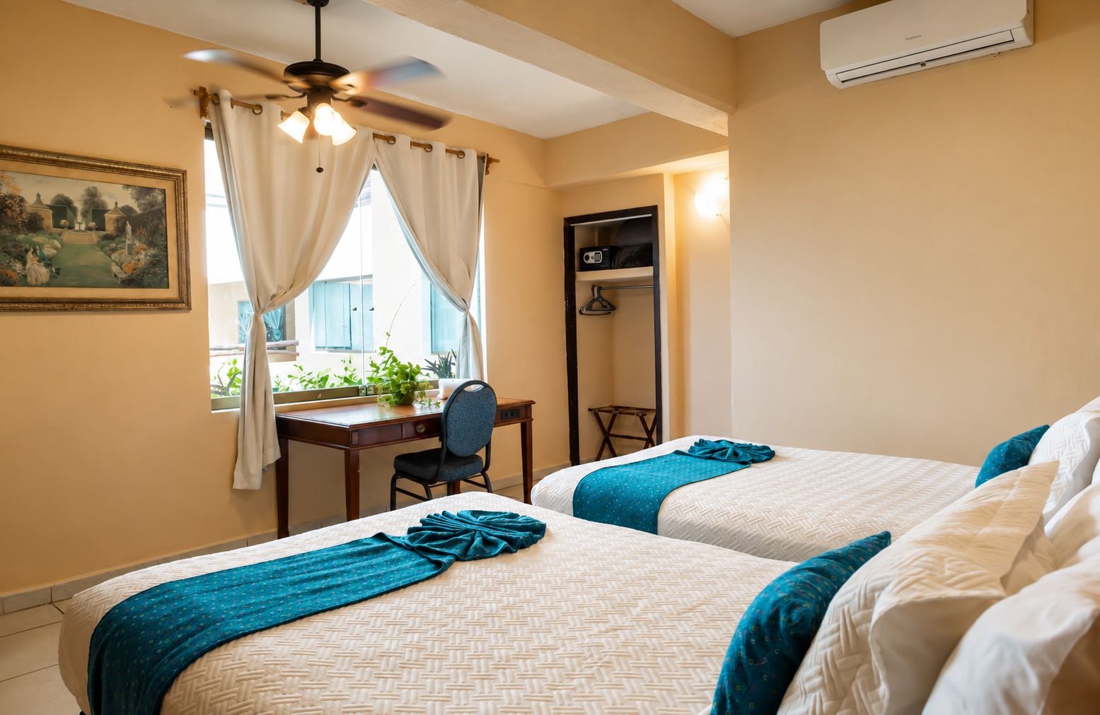 A hotel room with two beds, a desk, a chair, a ceiling fan, and a window with curtains.
