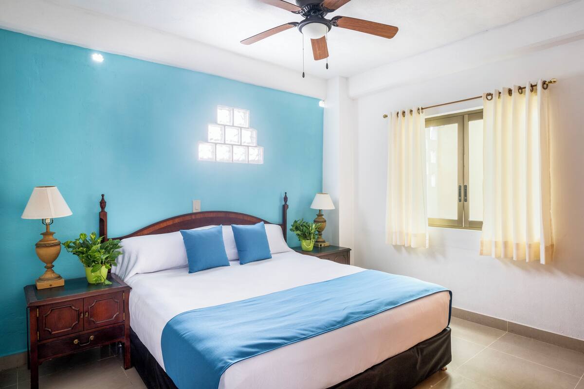 A bedroom with a bed, two nightstands, a ceiling fan, a window with curtains, and a wall clock.