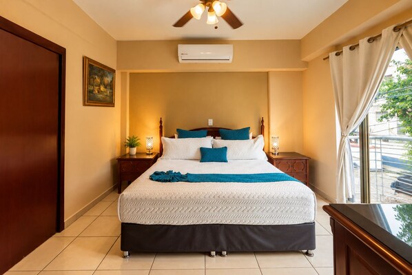 A hotel room with a bed, nightstands, an air conditioner, a ceiling fan, and a window with curtains.