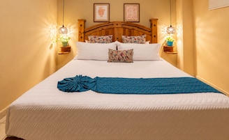 A bed with a quilted white cover, a blue blanket, and a decorative blue knot. There are two wall-mounted lamps on either side of the bed, and a wooden headboard.